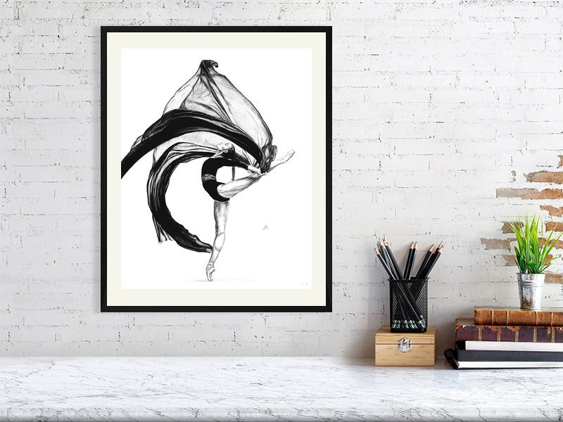 A large framed pencil drawing featuring a ballet dancer in a dynamic pose with flowing black gauze fabric. Artwork hangs in a black frame on a white brick wall over a desk.