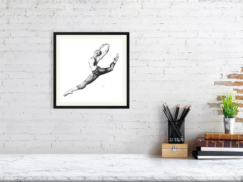 A medium size black framed pencil drawing of a ballet dancer in mid-leap, hanging on a white brick wall over a desk.