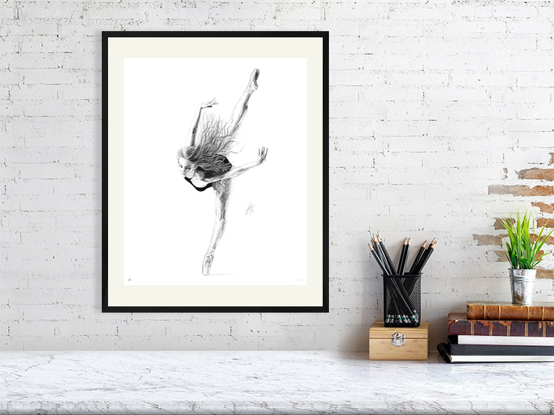 A large framed pencil drawing on a white background of a ballet dancer en pointe in mid-bend, with limbs outstretched and loose hair. Signed with Sophie Hall's monogram and hanging on a white brick wall over a desk.