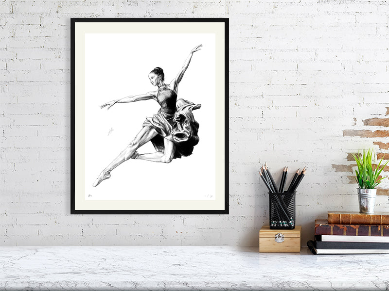 A large framed pencil drawing of a ballet dancer in mid-jump, by Sophie Hall, hanging on a white brick wall.