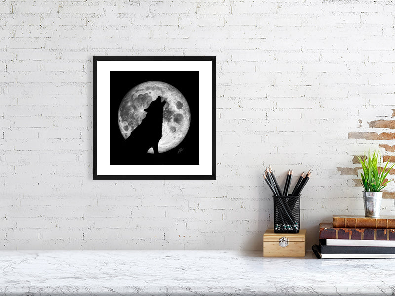 A black framed pencil drawing of a wolf howling at the moon, with the outline of the wolf silhouetted by the a large full moon behind, on a black paper background. Hanging on a white brick wall over a desk.