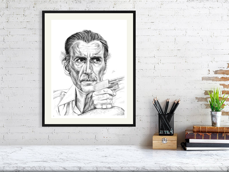A large framed pencil drawing of Christopher Lee as Antonin Scaramanga from The Man with the Golden Gun. Signed by Sophie Hall and hanging on a white brick wall over a desk.