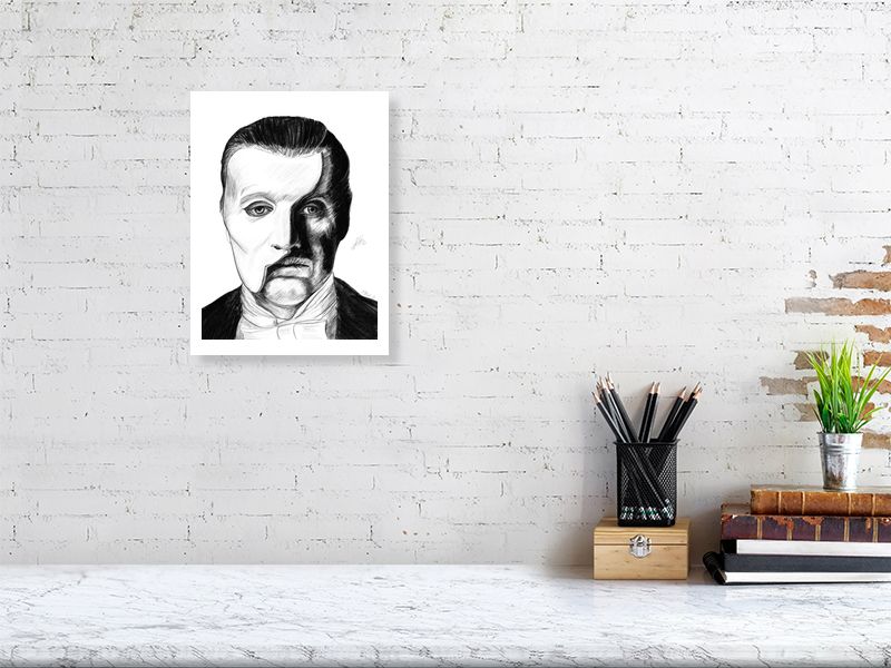 A medium unframed pencil drawing of the Phantom of the Opera, with a split portrait showing the character's face and iconic white half mask. Signed on the right by Sophie Hall and hanging on a brick wall over a desk.