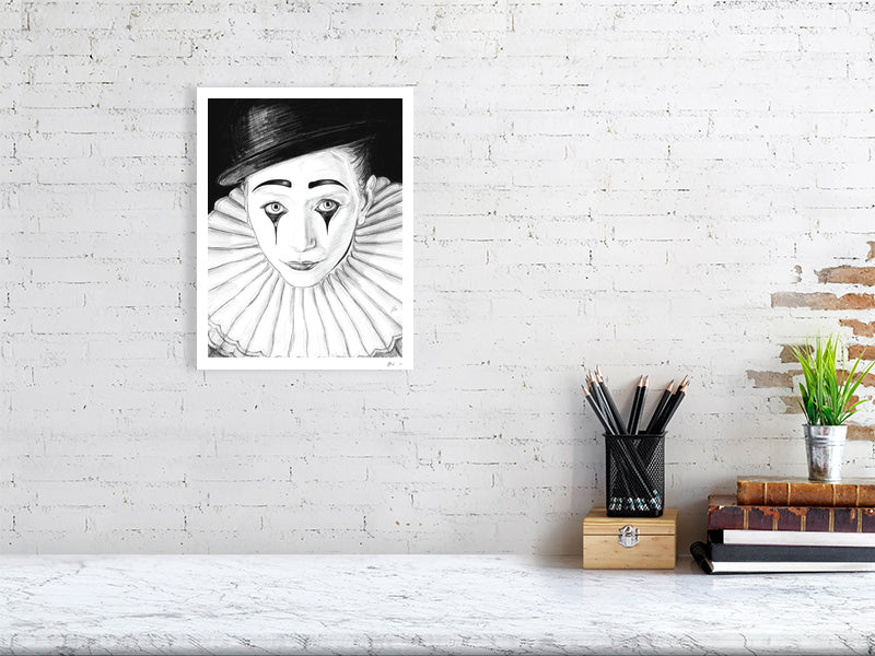 A medium size unframed pencil drawing of a sad Pierrot clown with white face paint, black hat, and large white collar. Picture hangs on a white brick wall over a desk.