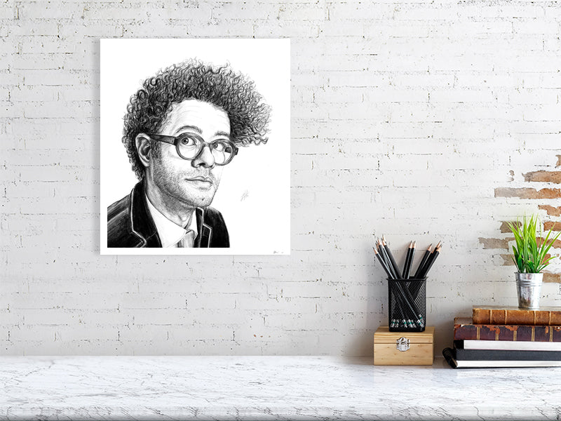 A large square unframed pencil drawing portrait of Richard Ayoade, hanging on a white brick wall over a marble desk.