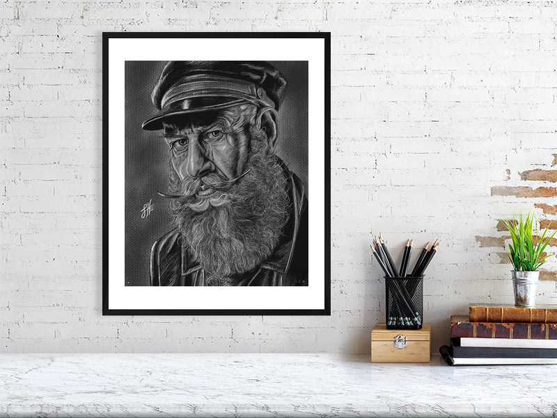 A large black and white framed pencil and chalk portrait drawing of an old sailor in monochrome with a grey background, hanging on a white brick wall over a desk.