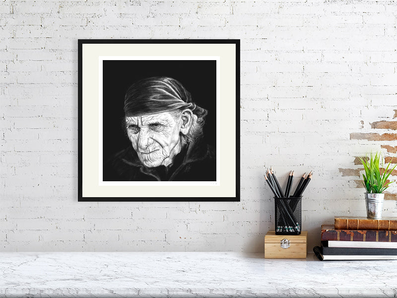 A large framed print monochrome pencil drawing portrait featuring a very wrinkled person with white wispy hair and a bandana, on a black background. Signed in the right corner SH '21 and hanging on a white brick wall over a desk.