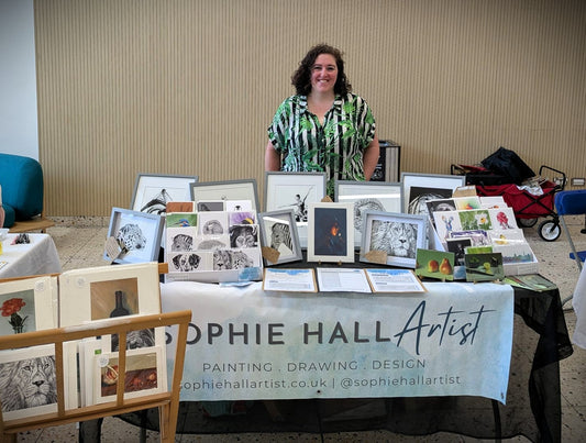 A person in a green and black patterned shirt stands behind an art fair stall featuring the artworks, prints and cards of Sophie Hall Artist