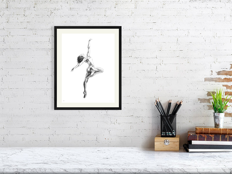 Medium framed pencil drawing of a ballet dancer mid Pique pose, with arms outstretched, signed in the corner by Sophie Hall and hanging on a white wall over a marble desk.