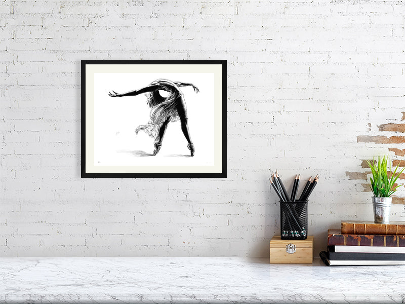 A framed print of a pencil drawing of a ballet dancer mid pose, by Sophie Hall. Print hangs on a white brick wall.
