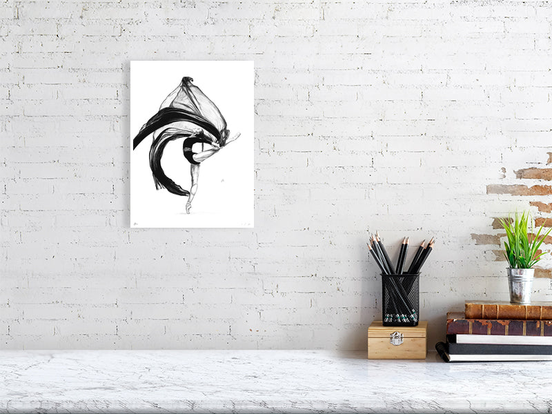 An unframed detailed pencil drawing featuring a ballet dancer in a dynamic pose with flowing black gauze fabric, hanging on a white brick wall over a desk.