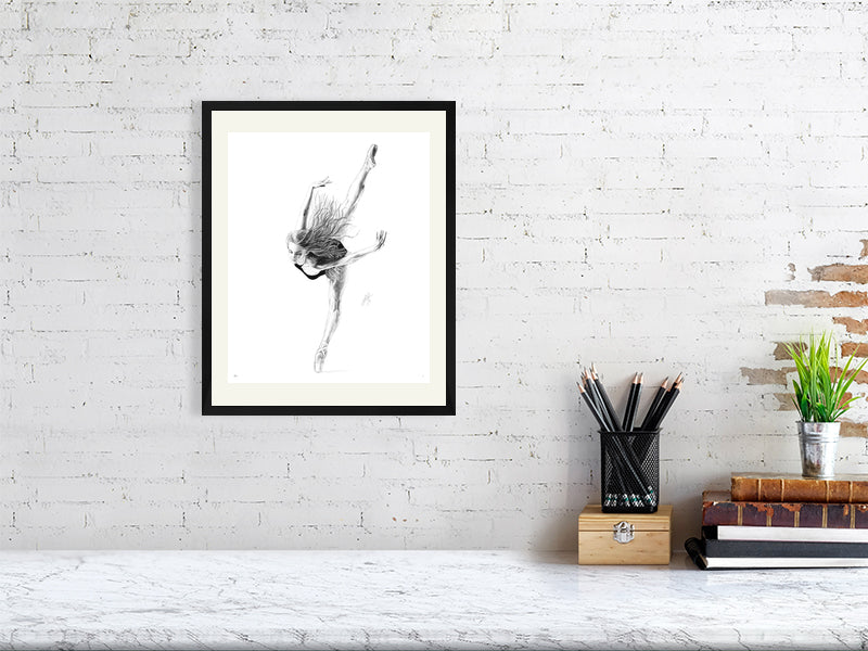 A black framed pencil drawing on a white background of a ballet dancer en pointe in mid-bend, with limbs outstretched and loose hair. Signed with Sophie Hall's monogram and hanging on a white brick wall over a desk.