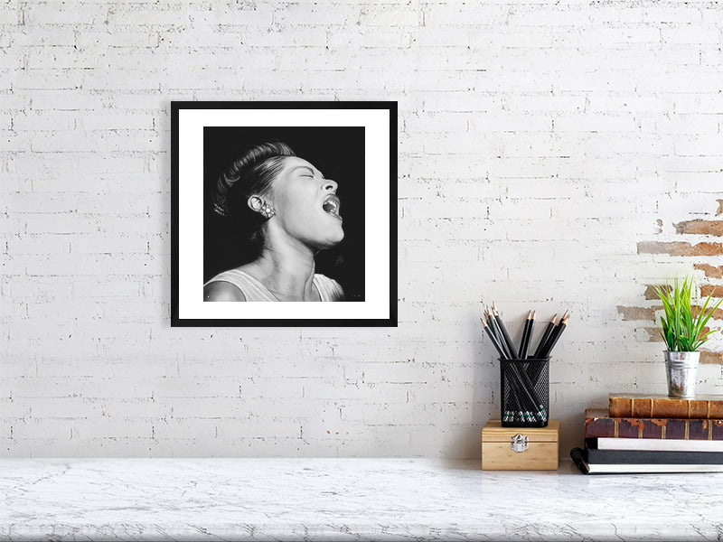 A medium framed black and white portrait pencil drawing of Billie Holiday, depicting the singer with a expressive pose and closed eyes, capturing a sense of emotion and intensity. Hanging on a white brick wall over a desk.