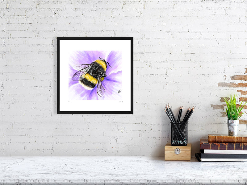 A medium framed watercolour and pencil artwork of a fluffy yellow bumblebee on a purple flower, signed by Sophie Hall and hanging on a white brick wall over a desk.