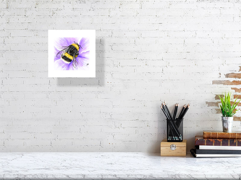 A small unframed watercolour realistic artwork of a fluffy yellow bumblebee on a purple flower, signed by Sophie Hall and hanging on a white brick wall over a desk.