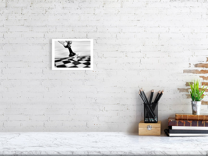 A small unframed pencil drawing of a chess piece moving on a checkered board to knock another piece over, hanging on a white brick wall over a desk.