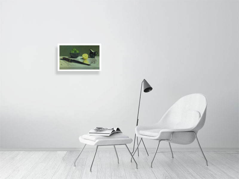 Small unframed print of traditional style oil painting featuring citrus fruits in a bowl, a lemon cut in half, a black jug, and a knife placed on a reflective marble surface. Hanging on a white wall above a white chair and lamp.