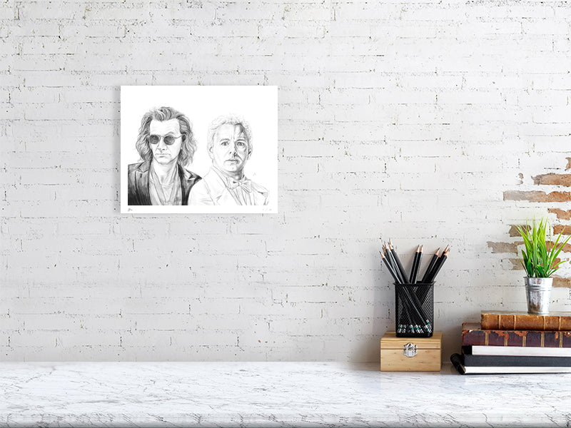 A small unframed detailed pencil drawing of the characters Crowley and Aziraphale from the TV show Good Omens, signed by artist Sophie Hall and hanging on a white wall over a desk.