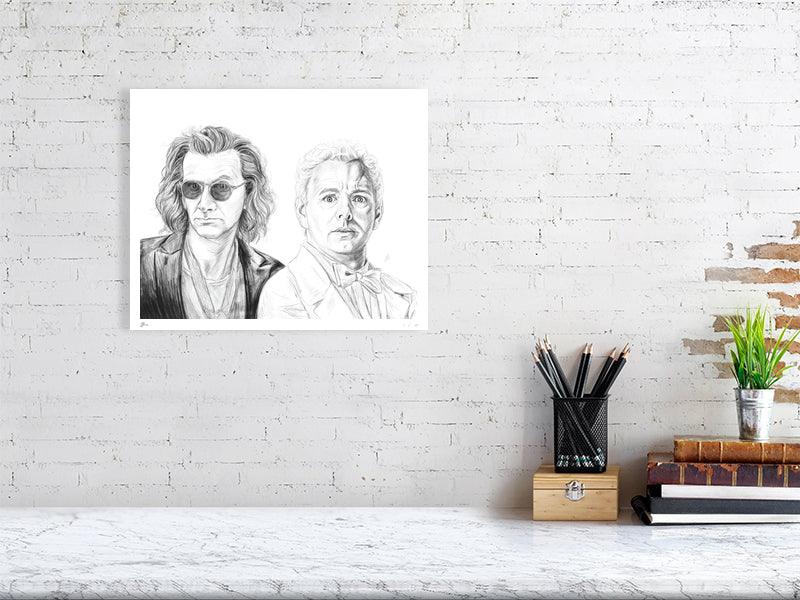 A large unframed detailed pencil drawing of the characters Crowley and Aziraphale from the TV show Good Omens, signed by artist Sophie Hall and hanging on a white wall over a desk.