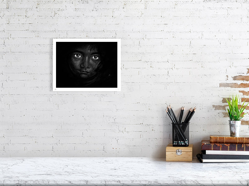 A medium unframed chalk and pencil portrait drawing on a dark background of a young child with striking emotive eyes, signed in the corner by Sophie Hall and hanging on a white brick wall.