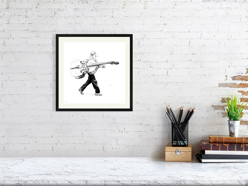 A medium framed pencil drawing of a man in a flat cap carrying a large bass instrument under one arm and striding towards the right. Artwork hangs on a white brick wall over a desk.