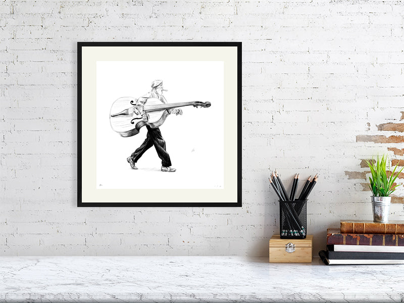 A large framed pencil drawing of a man in a flat cap carrying a large bass instrument under one arm and striding towards the right. Artwork hangs on a white brick wall over a desk and is framed with a cream mount and black wood.
