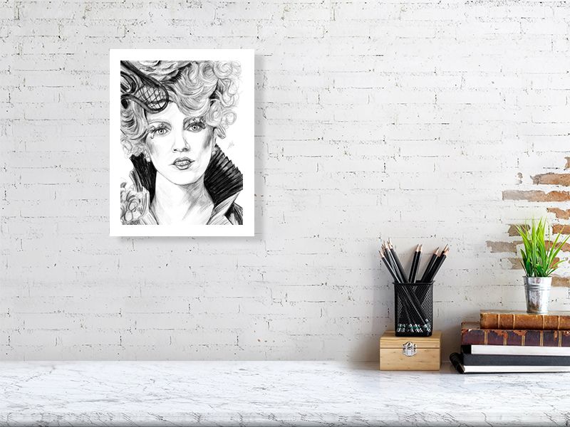 An unframed detailed monochrome pencil portrait of the character Effie Trinket from The Hunger Games, signed by Sophie Hall and hanging on a white brick wall.