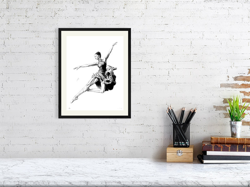 A medium size framed pencil drawing of a ballet dancer in mid-jump, by Sophie Hall, hanging on a white brick wall.