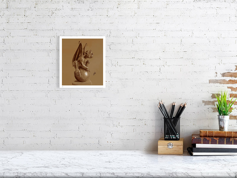 A small unframed pencil drawing of a winged gargoyle creature sitting on a ball, rendered in brown pencil and chalk on a textured sienna paper and hanging on a white brick wall over a desk.