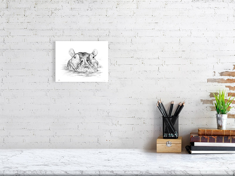 A small unframed print of a pencil drawing portrait of a hippo partially submerged in water, signed by Sophie Hall and hanging on a white brick wall above a desk.