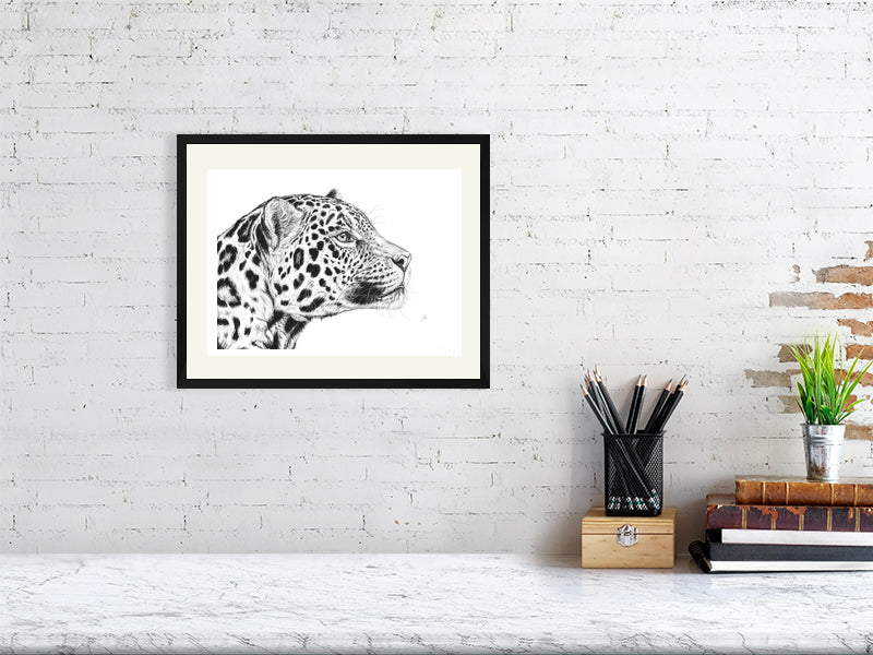 A small framed pencil portrait drawing of a jaguar with a detailed and lifelike depiction, hanging on a white brick wall above a desk.