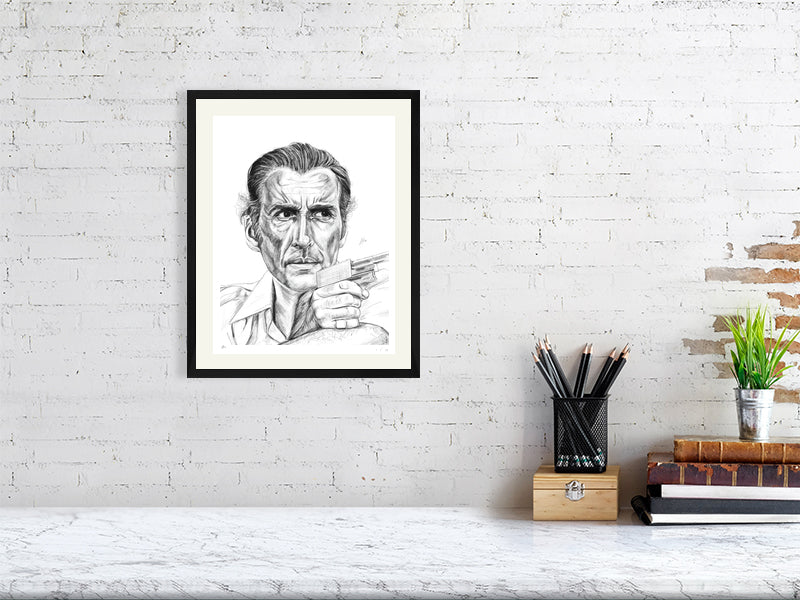 A medium size framed pencil drawing of Christopher Lee as Antonin Scaramanga from The Man with the Golden Gun. Signed by Sophie Hall and hanging on a white brick wall over a desk.
