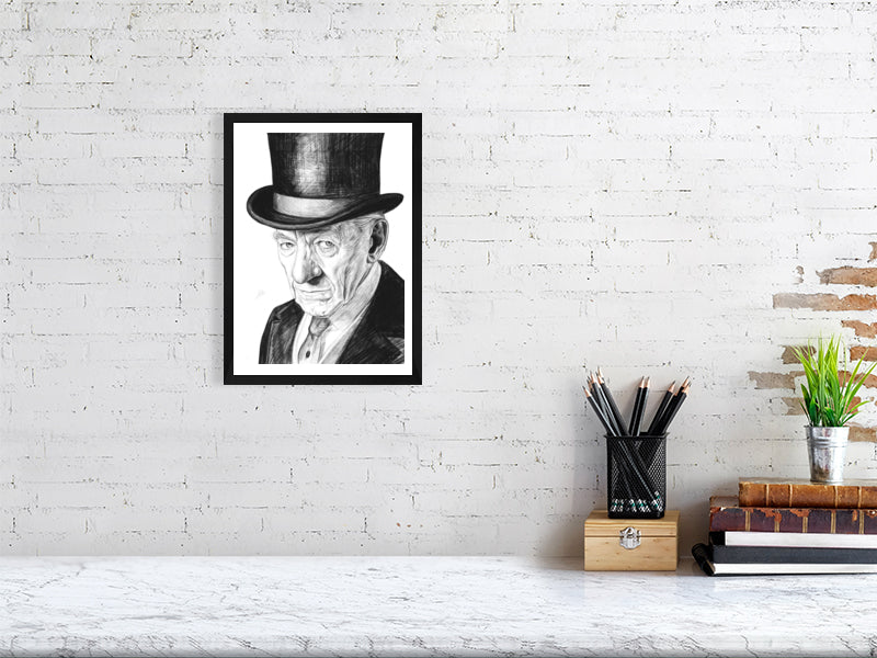 A medium size black framed pencil drawing of Ian McKellen wearing a black top hat as Sherlock Holmes. Signed with Sophie Hall's monogram and hanging on a white brick wall above a desk.