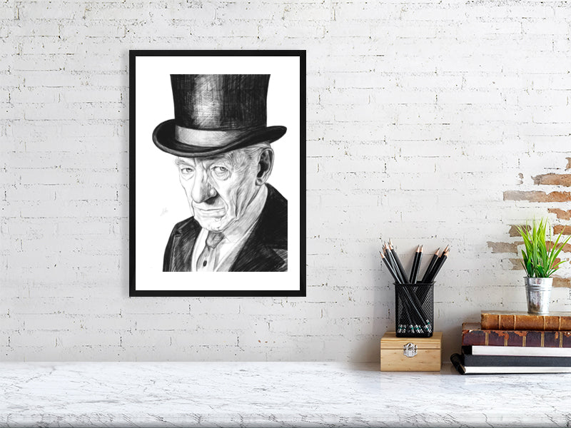 A large black framed pencil drawing of Ian McKellen wearing a black top hat as Sherlock Holmes. Signed with Sophie Hall's monogram and hanging on a white brick wall above a desk.