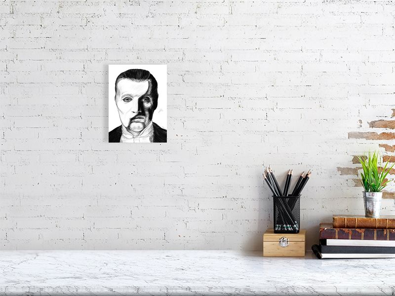 A small unframed pencil drawing of the Phantom of the Opera, with a split portrait showing the character's face and iconic white half mask. Signed on the right by Sophie Hall and hanging on a brick wall over a desk.