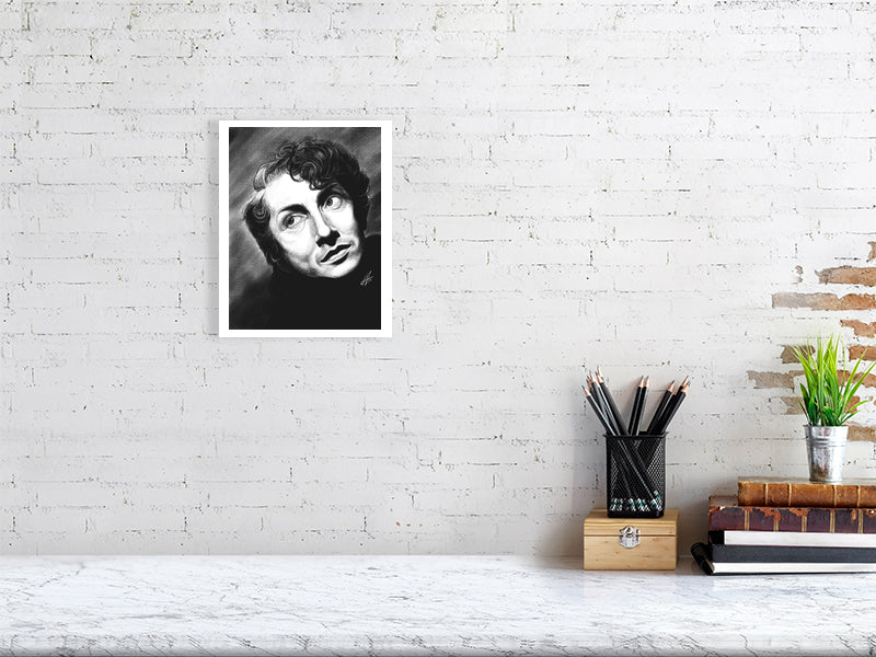 An unframed black and white pencil drawing of Quentin Crisp, featuring expressive facial features and a direct gaze. Hanging on a white brick wall over a desk.