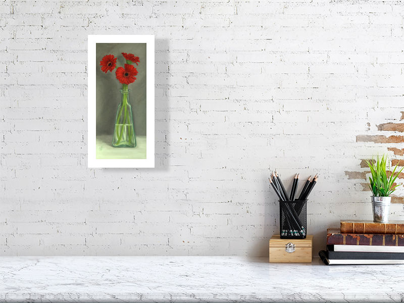 Small unframed print of traditional style oil painting featuring three bright red gerbera flowers in a clear glass bottle, with a grey background. Signed by the artist Sophie Hall and hanging on a white brick wall over a desk.