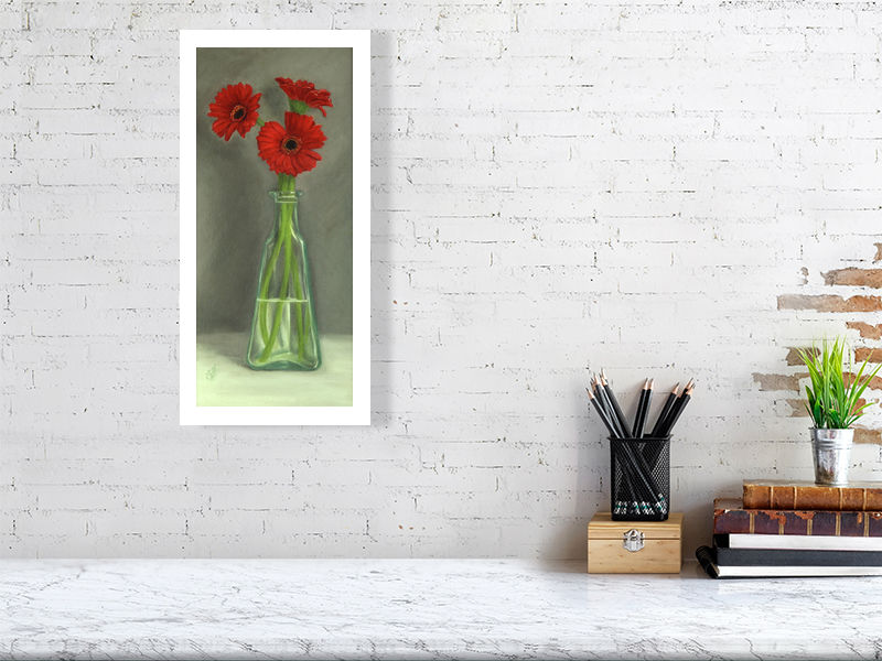 Large unframed print of traditional style oil painting featuring three bright red gerbera flowers in a clear glass bottle, with a grey background. Signed by the artist Sophie Hall and hanging on a white brick wall over a desk.