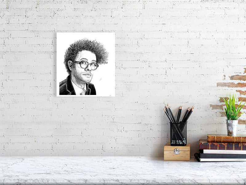 A medium unframed pencil drawing portrait of Richard Ayoade, hanging on a white brick wall over a marble desk.