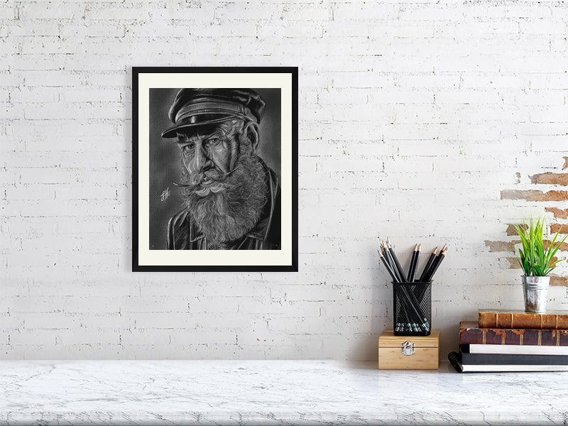 A black and white framed pencil and chalk portrait drawing of an old sailor in monochrome with a grey background, hanging on a white brick wall over a desk.