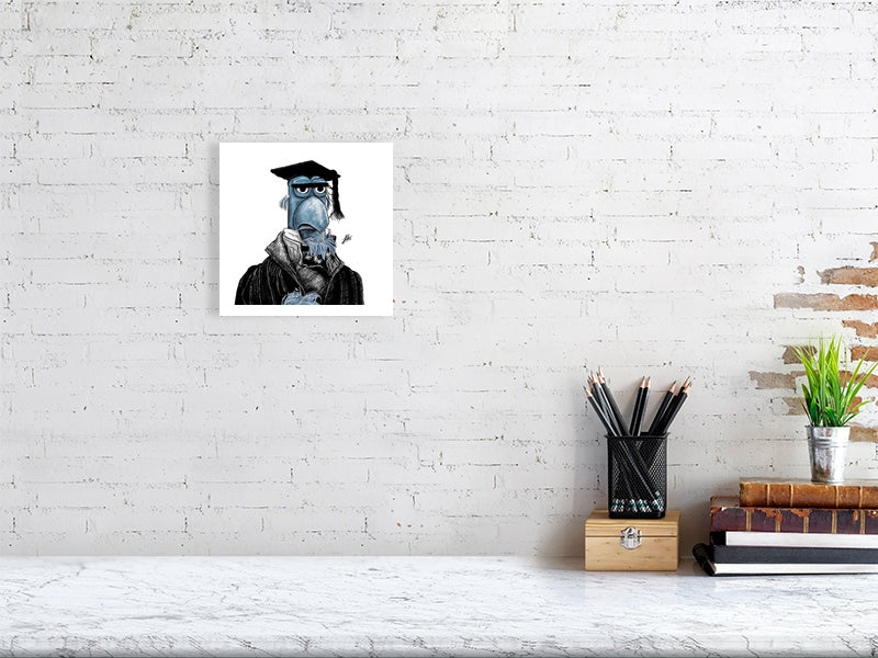 A small unframed print hanging on a white brick wall above a desk. The artwork features a detailed ink drawing of Sam the blue Eagle from the Muppets Christmas Carol, wearing a black coat, white shirt, and black teacher mortar board hat. Monogram signed by Sophie Hall.