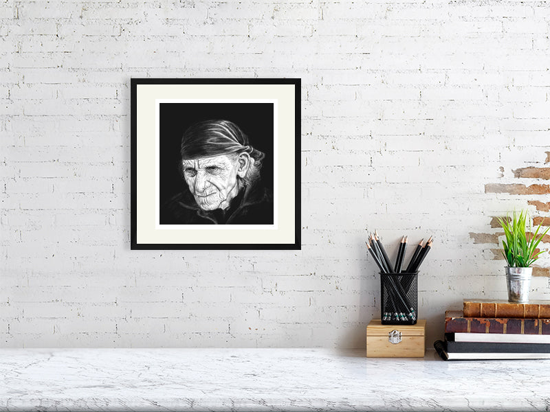A framed print monochrome pencil drawing portrait featuring a very wrinkled person with white wispy hair and a bandana, on a black background. Signed in the right corner SH '21 and hanging on a white brick wall over a desk.