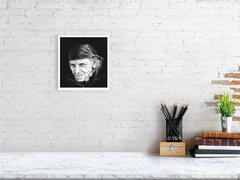 A small unframed print of a highly detailed monochrome pencil drawing portrait, featuring a very wrinkled person with white wispy hair and a bandana, on a black background. Signed in the right corner SH '21 and hanging on a white brick wall over a desk.