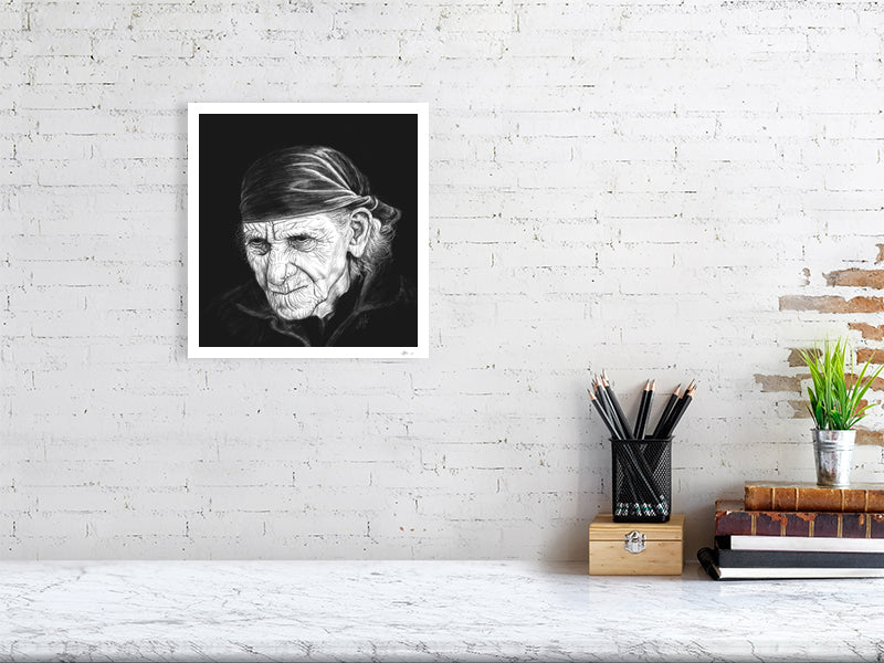 A medium square unframed print of a highly detailed monochrome pencil drawing portrait, featuring a very wrinkled person with white wispy hair and a bandana, on a black background. Signed in the right corner SH '21 and hanging on a white brick wall over a desk.