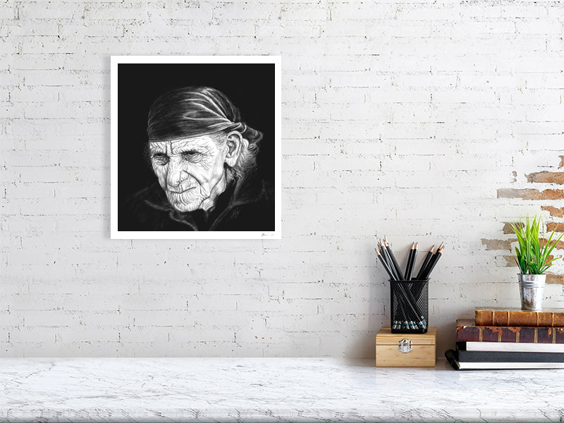 A large unframed print of a highly detailed monochrome pencil drawing portrait, featuring a very wrinkled person with white wispy hair and a bandana, on a black background. Signed in the right corner SH '21 and hanging on a white brick wall over a desk.