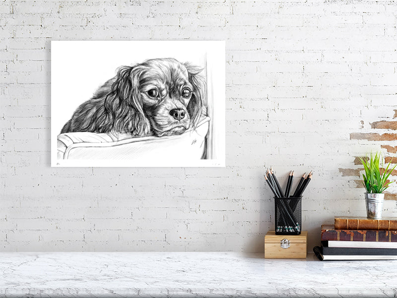 A large unframed pencil drawing of a sad-looking spaniel with long ears, perching its head over the back of a chair. Print hangs on a white brick wall above a desk and is signed by Sophie Hall.