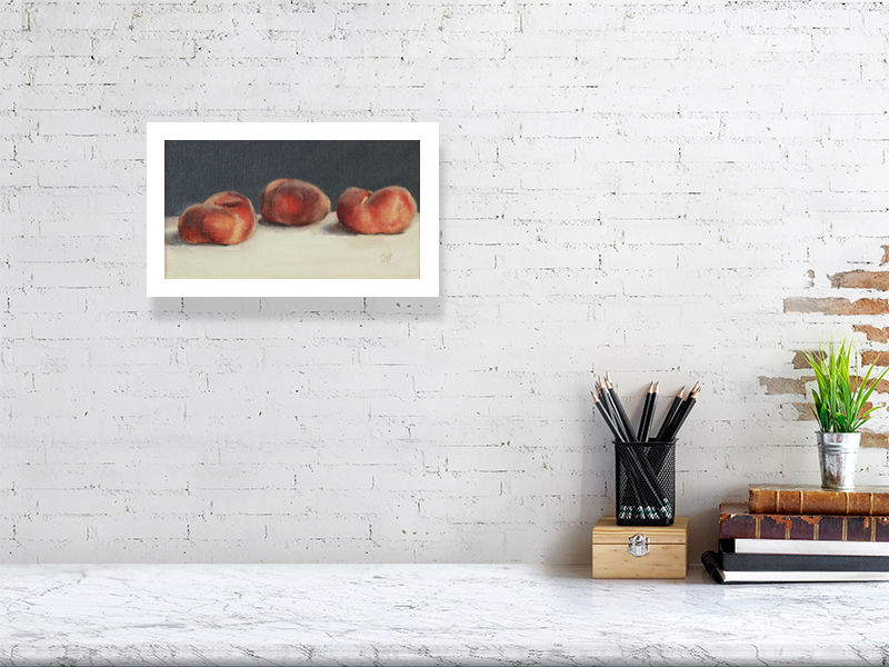 A small unframed print of Three Flat Peaches, a traditional still life oil painting of three flat peaches on a white piece of linen, signed by Sophie Hall. The print hangs on a white brick wall above a desk.