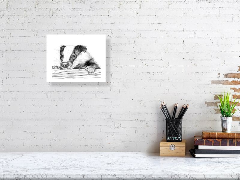 A medium unframed pencil drawing portrait of a baby badger, peeking over a basket edge. By Sophie Hall. Print hangs on a white brick wall.