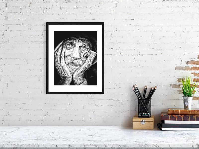 A medium framed pencil portrait depicting an elderly man with his chin in his hands and a contemplative expression. Print hangs in a black wood frame on a white brick wall above a desk.