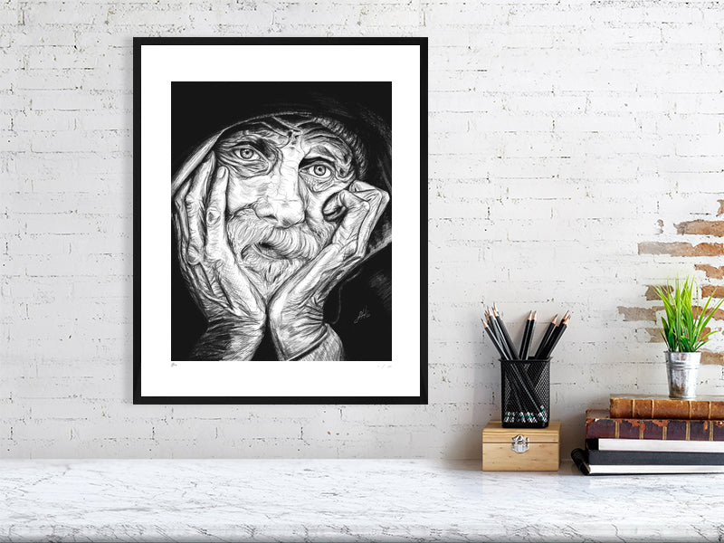 A large framed pencil portrait depicting an elderly man with his chin in his hands and a contemplative expression. Print hangs in a white mount and black wood frame on a white brick wall above a desk.
